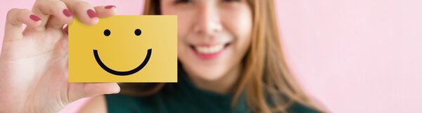 Eine lächelnde Frau mit langen Haaren hält eine gelbe Karte mit einem schwarzen Smiley-Gesicht vor ihr Gesicht; Hintergrund ist rosa.